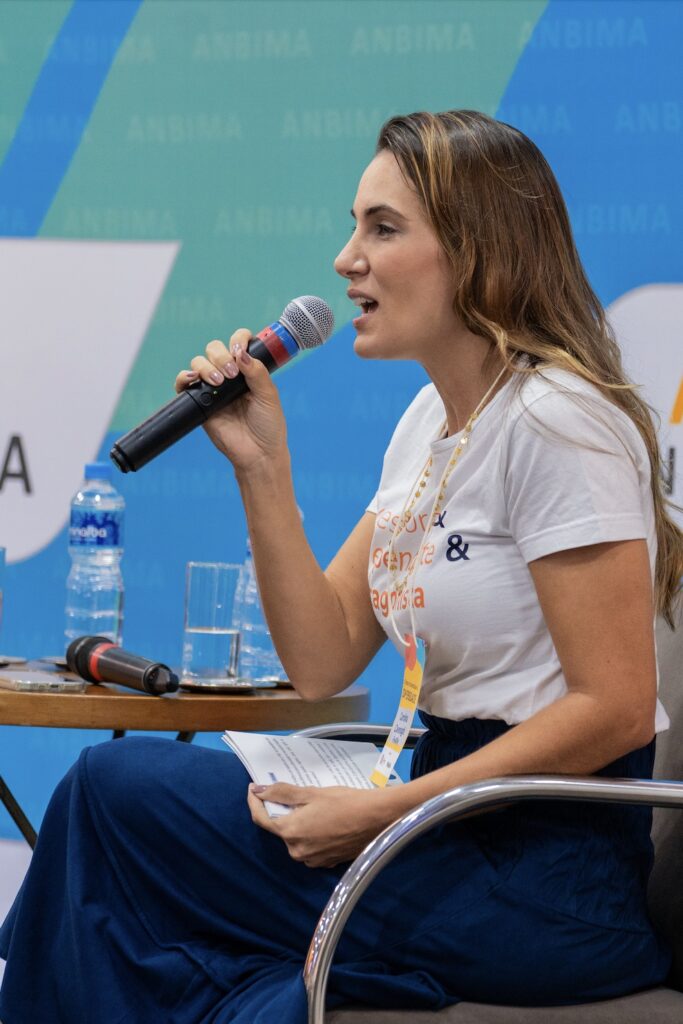 Fotografia de Carolina Cavenaghi, uma mulher branca, de cabelos loiros lisos na altura da lombar. Ela está sentada em uma cadeira em um palco, segura um microfone e é fotografada de lado enquanto fala.