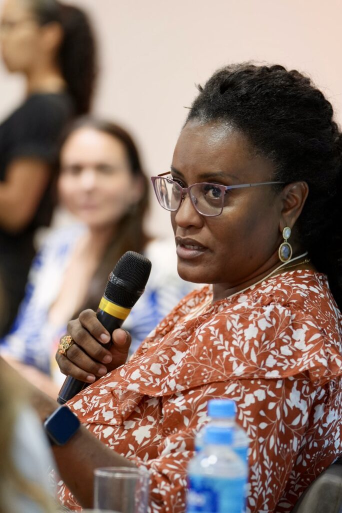 Fotografia de Jandaraci Araujo de perfil. Ela é uma mulher negra, de cabelos crespos longos, veste um vestido laranja e segura um microfone bastão enquanto fala.