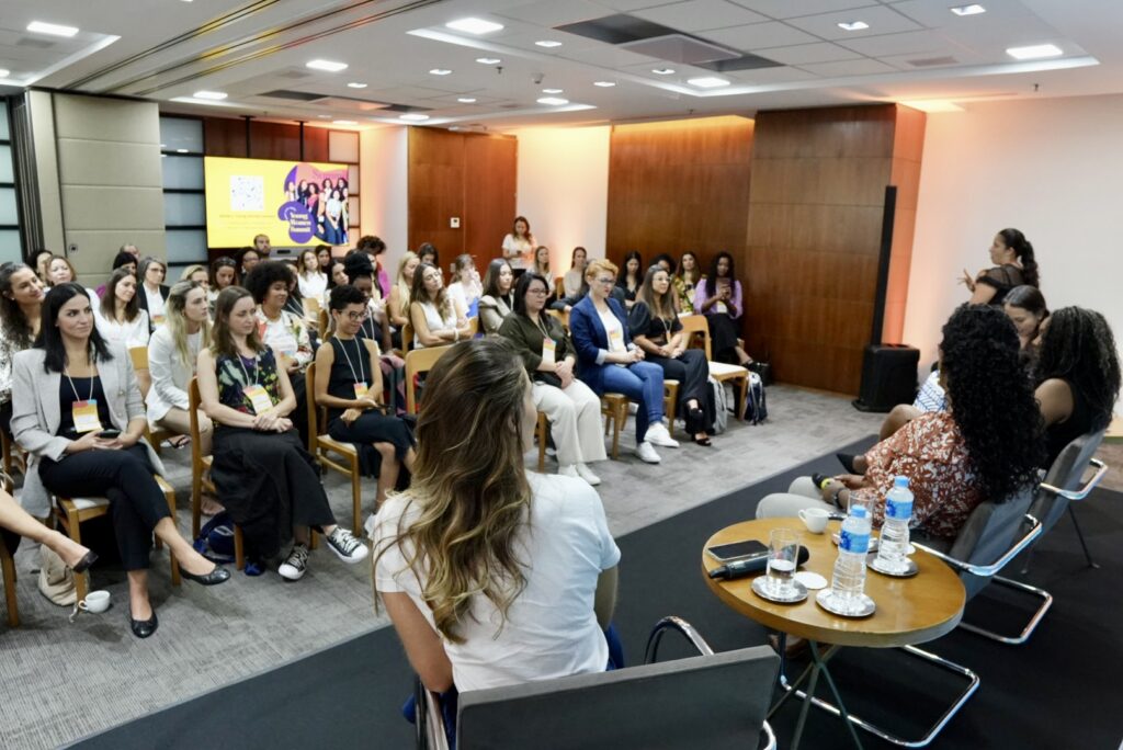 Fotografia exibe uma sala de escritório com dezenas de mulheres sentadas em cadeiras de madeira e olhando para o palco. Nele, há quatro mulheres sentadas de costas para a foto.