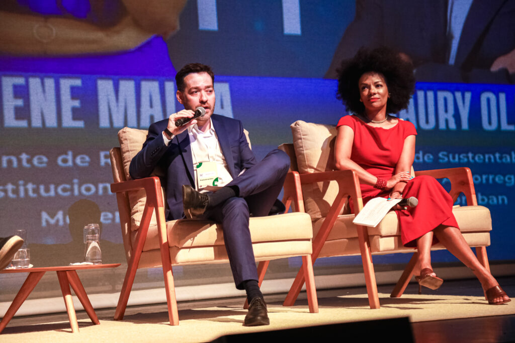 Fotografia de Amaury Oliveira no palco da Conferência Empresarial ESG Racial. Ele está sentado em uma poltrona e fala segurando um microfone bastão. Amaury é um homem branco, de cabelos castanhos curtos e veste um terno azul marinho com camisa branca.